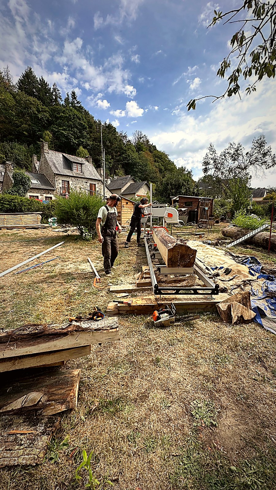Vue large du chantier de sciage mobile avec plots empilés et maisons en pierre bretonnes